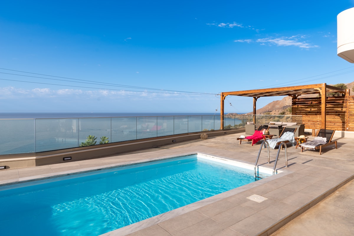 An outdoor area is highlighted by a private pool with steps leading down into the water. Lounge chairs are positioned around the pool, while a wooden pergola offers partial shade. The sea and mountains create a scenic backdrop, and the clear blue sky enhances the peaceful ambiance.