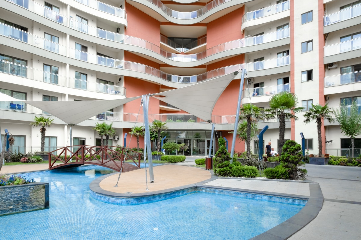 A spacious pool area is surrounded by palm trees and decorative plants, offering a serene setting. A wooden bridge crosses over a shallow section of water, while modern lounge chairs are positioned nearby. The architecture of the building features multiple levels with balconies overlooking the courtyard.