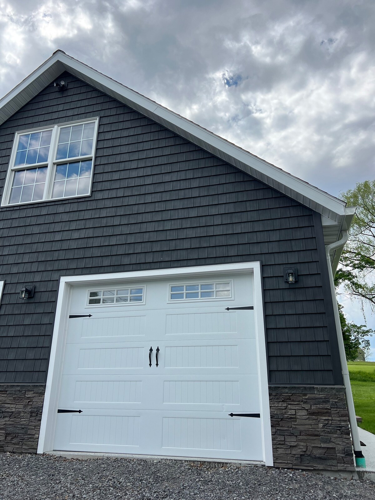 The exterior of a new home is shown, featuring a gray shingle facade complemented by a white garage door. Above the garage door, a large window allows natural light inside. Stone accents add texture to the lower part of the structure, under a partly cloudy sky.
