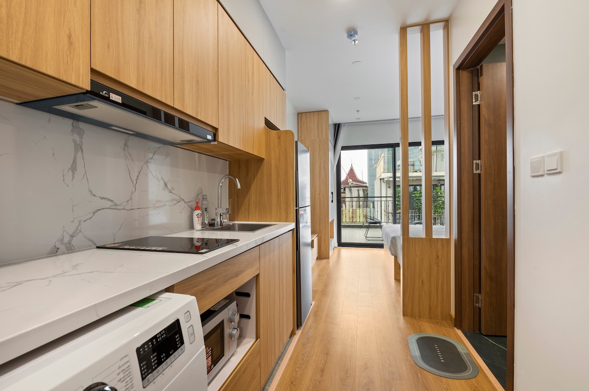 The kitchen features modern wooden cabinetry with a sleek countertop, including an integrated sink and stovetop. A refrigerator and microwave are visible, complemented by natural light streaming in through the glass doors leading to a balcony. The open layout extends towards a cozy sleeping area in the background.