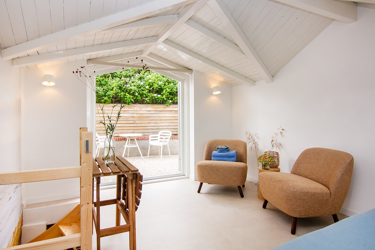The interior space features a sloped white ceiling with natural light flooding in through a large window. Two comfortable armchairs are positioned beside a small wooden table, while a staircase made of wood leads down, enhancing the airy atmosphere.