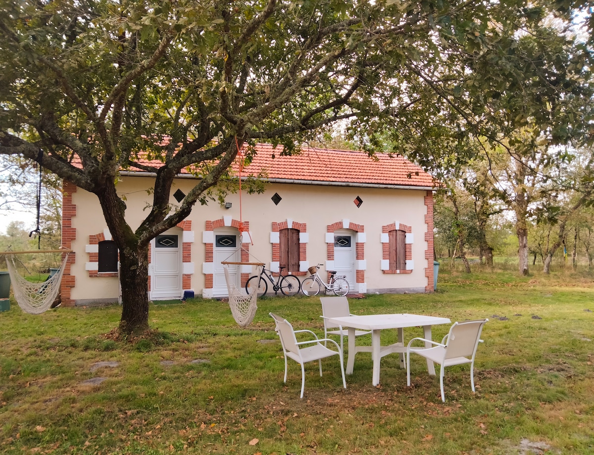 A charming building is set among trees, featuring a red-tiled roof and a light-colored exterior with contrasting brick accents. A small table with four chairs is situated outside, with two bicycles leaning against the wall. Hammocks are draped from the trees, providing shaded relaxation.