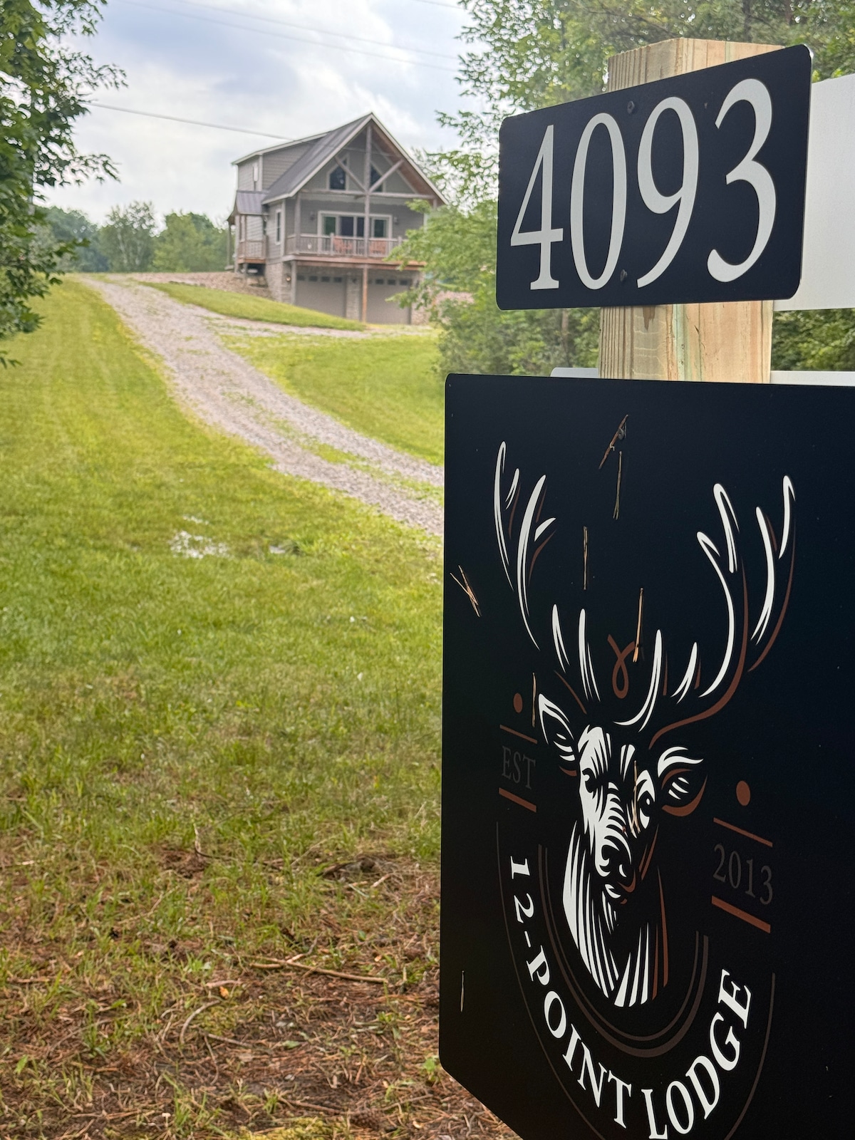 A welcoming lodge sign featuring a deer design is positioned beside a gravel driveway. The cabin, visible in the distance, is framed by lush greenery and trees, set against a gently rolling landscape. The property is identified by the address 4093.
