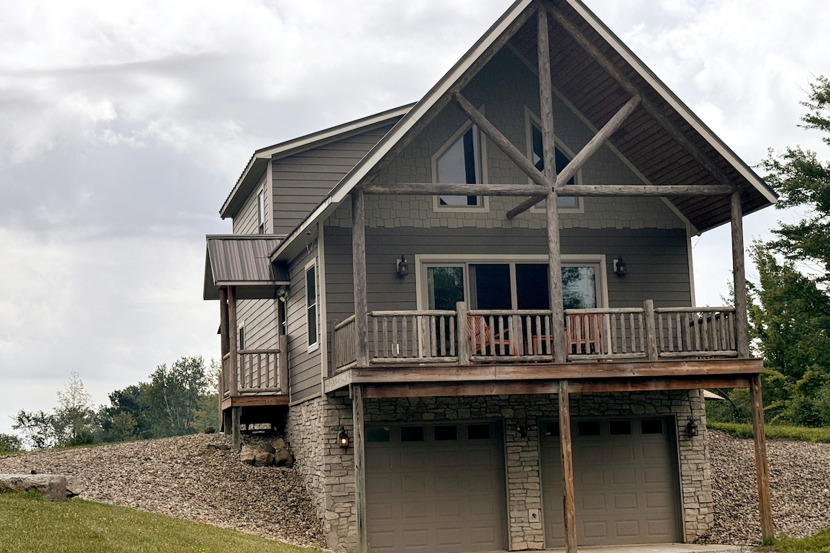 The exterior of a two-story cabin is presented, featuring a combination of stone and wood elements. A covered porch with wooden railings overlooks the landscape, while large windows enhance natural light. Two garage doors are visible beneath the structure, set against a rocky slope.