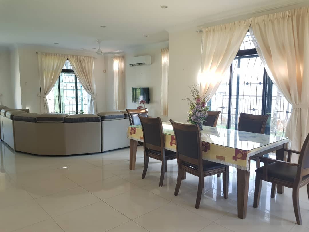 A bright and open living area features a large sectional sofa and a dining table set for eight guests. Natural light filters through floor-to-ceiling windows adorned with sheer curtains, enhancing the spacious ambiance. Air conditioning is available for comfort.
