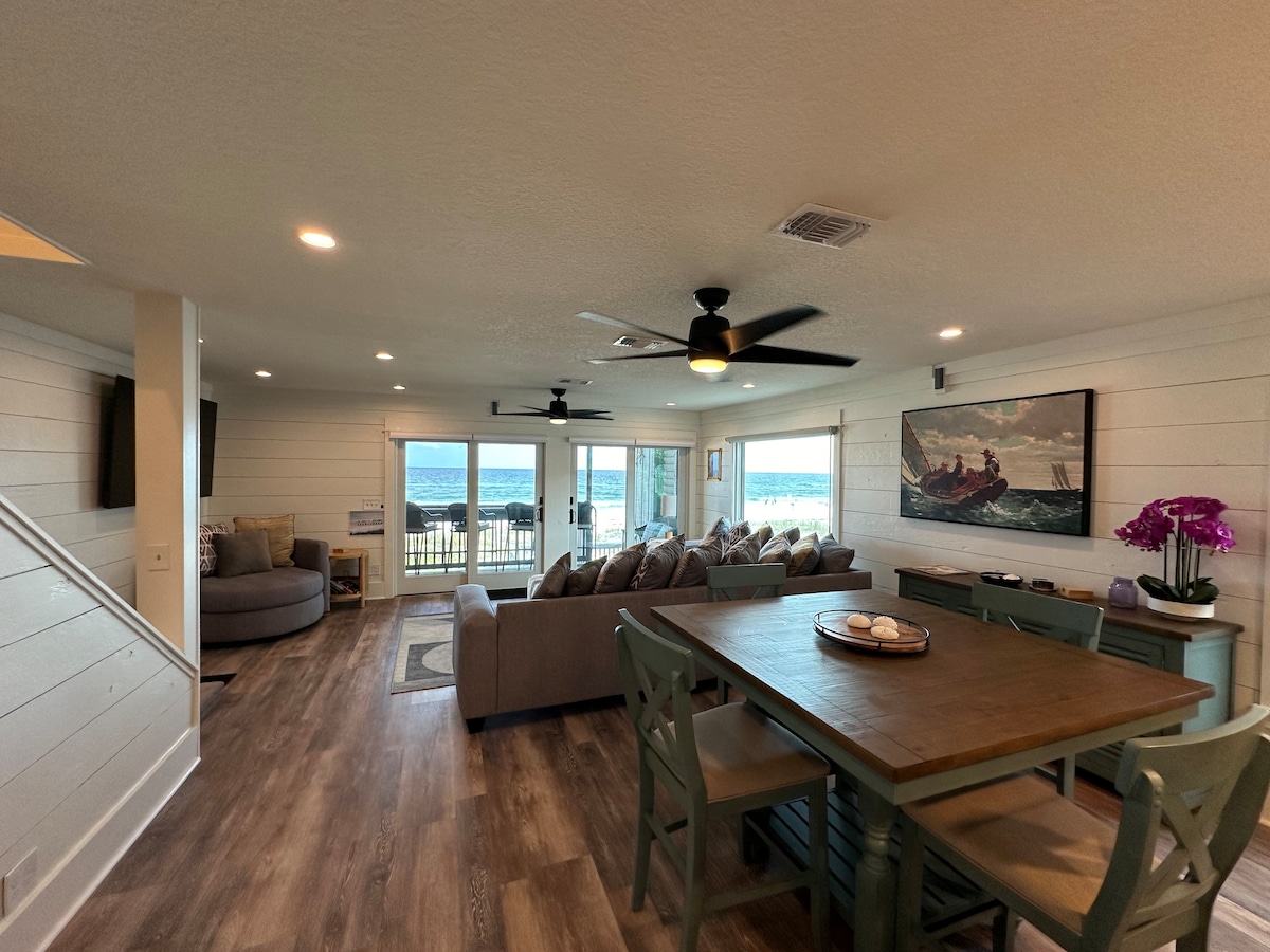 An open living area features a large sectional sofa arranged around a coffee table. A dining table with wooden chairs is positioned nearby. Large glass doors provide abundant natural light and showcase views of the ocean. Modern decor is complemented by a wall-mounted television.