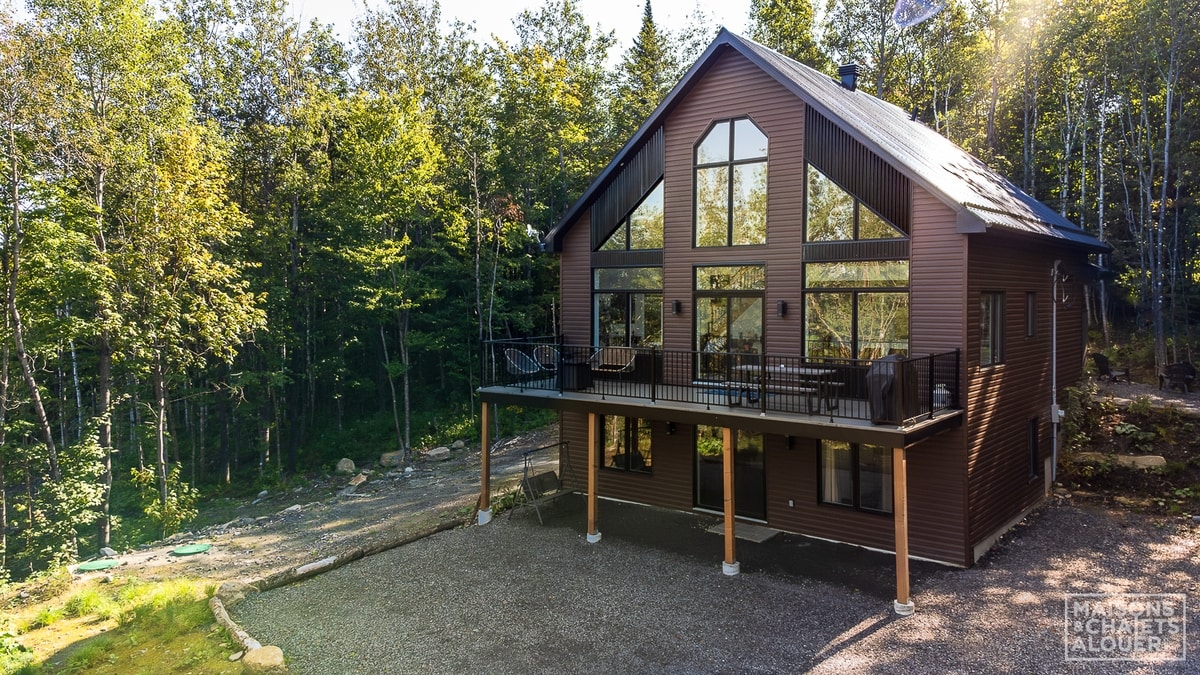 A modern chalet is set against a backdrop of lush trees. Large glass windows dominate the upper floor, allowing natural light to flood the interior. A spacious balcony extends from the front, furnished for relaxation, while the ground level is accessible through a gravel path.