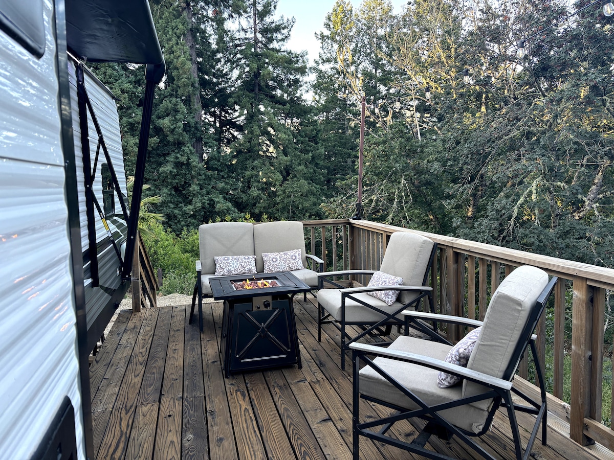 A spacious deck features a seating arrangement with four light-colored chairs surrounding a black fire table. Surrounding lush greenery provides a natural backdrop, highlighting a peaceful outdoor space designed for relaxation.