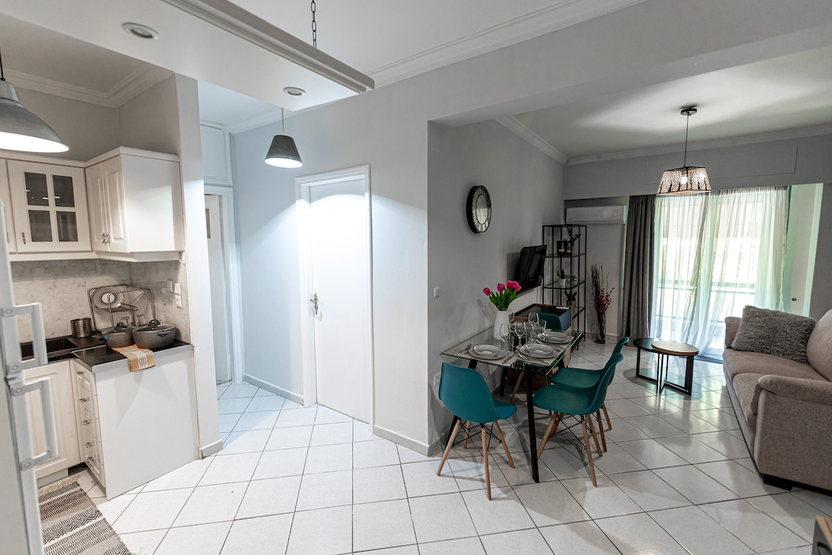 An open-plan living area features a dining table set for four, surrounded by teal chairs. A comfortable sofa is positioned nearby, and natural light fills the space through large windows. The adjacent kitchen includes modern cabinetry and appliances, enhancing the functional design.