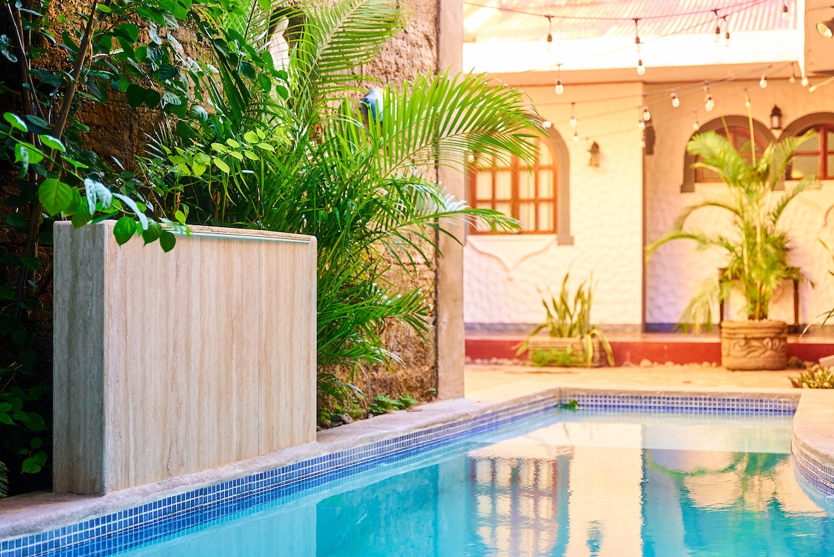 A clear swimming pool reflects the surrounding greenery and ambient lighting. A stone water feature adds a serene element, while well-maintained tropical plants frame the area, enhancing the inviting courtyard atmosphere.