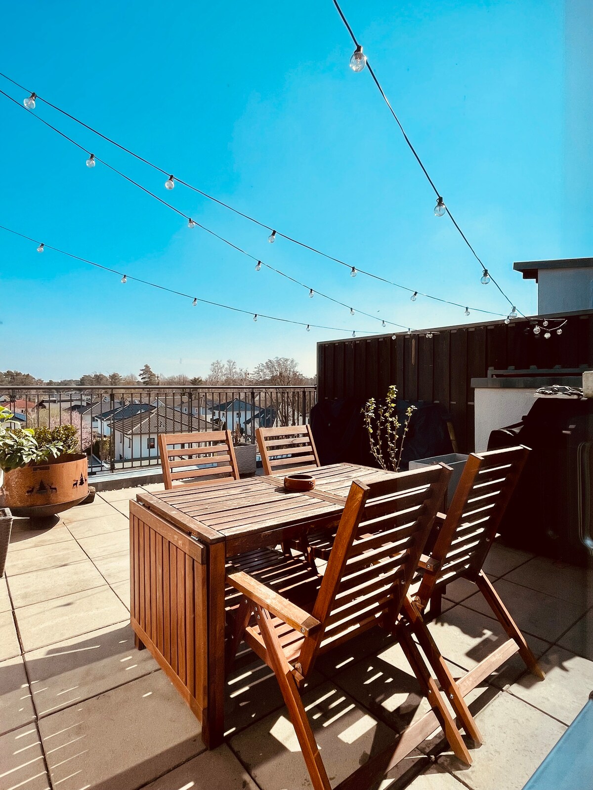 An outdoor terrace features a wooden dining table surrounded by folding chairs, illuminated by string lights overhead. The space is complemented by potted plants, with a clear blue sky in the background, offering a serene environment for relaxation.