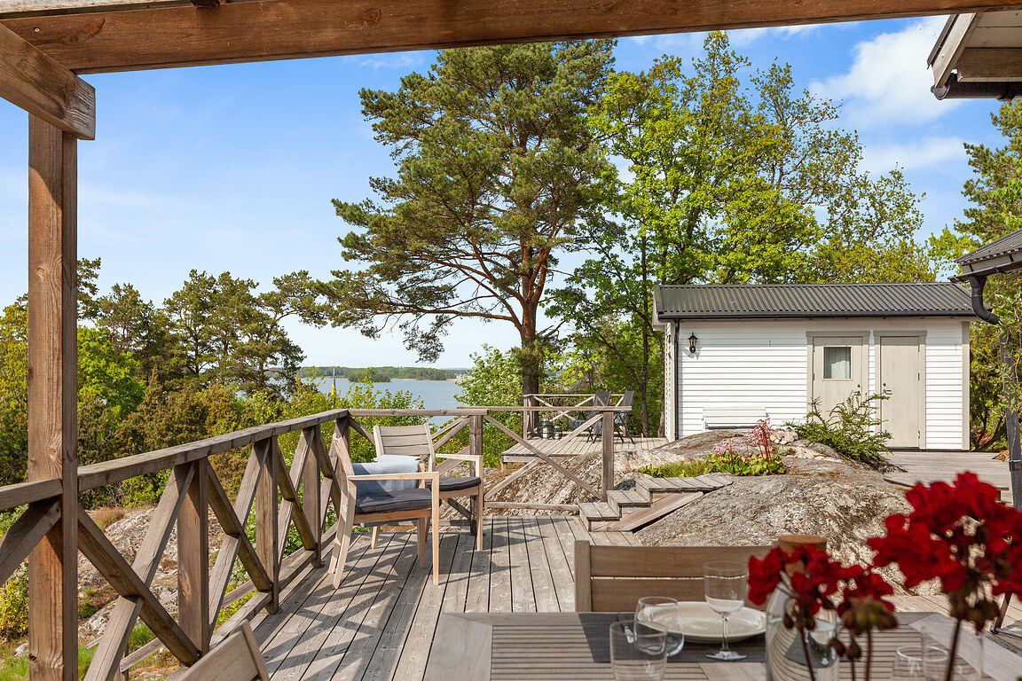 A spacious wooden terrace presents a scenic view overlooking water and surrounding greenery. A white guest cabin is visible in the background, complementing the natural landscape. Seating areas are arranged for relaxation, framed by the wooden railing and decorated with potted plants.