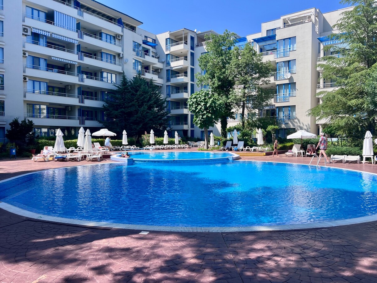 A large swimming pool is surrounded by comfortable lounge chairs and umbrellas. Lush greenery is visible around the pool area, providing shade. Several multi-story buildings are situated in the background, suggesting a vibrant community atmosphere. Pathways connect different sections of the poolside area.