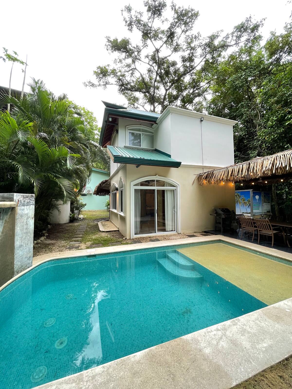 A two-story building with a green roof is presented beside a swimming pool. Palm trees and lush foliage surround the property, providing a natural feel. The pool area features a thatched-roof outdoor space with dining options, inviting relaxation and outdoor enjoyment.