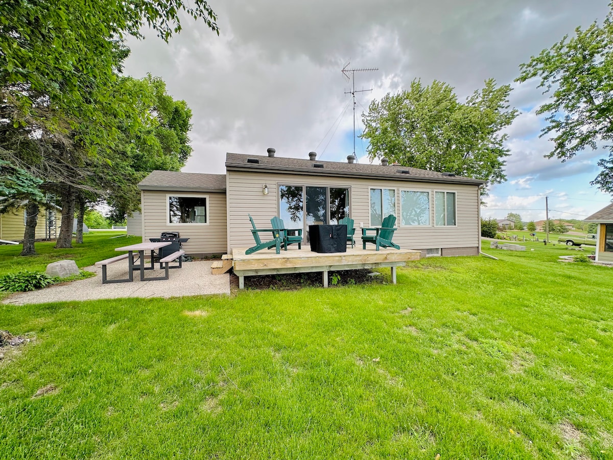 The cabin exterior showcases a welcoming lakefront design, featuring a spacious deck with outdoor chairs for relaxation. Lush green grass surrounds the property, complementing the tall trees and serene landscape. Large windows offer views into the interior living space.