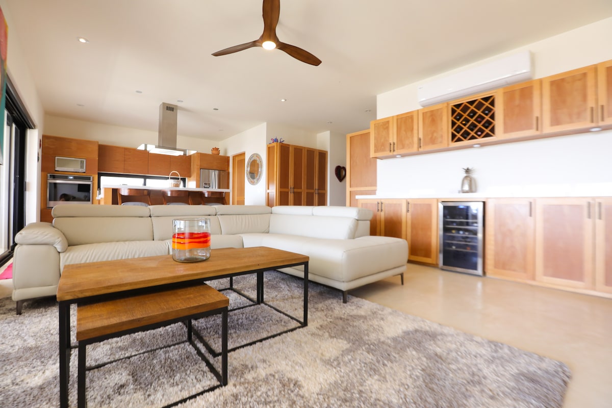 A spacious living area features a modern sectional sofa positioned near a wooden coffee table, complemented by a soft rug. Light wood cabinetry is visible in the background, along with stainless steel appliances. A ceiling fan circulates air above, highlighting the open layout.