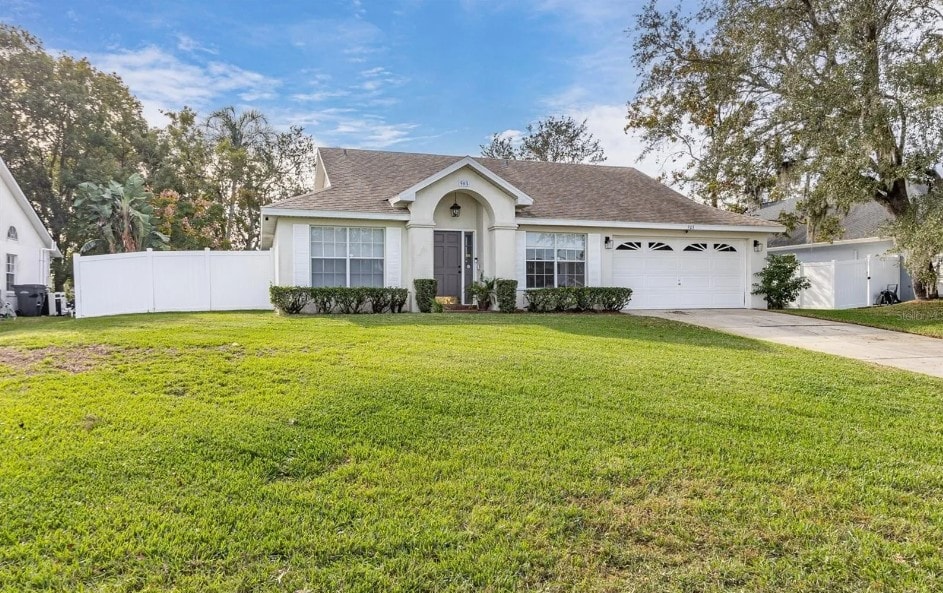 A single-story home showcases a well-maintained front lawn with lush green grass. The house features a welcoming entrance with a central door, flanked by large windows. A driveway leads to a two-car garage with decorative doors, while a fenced yard adds privacy.