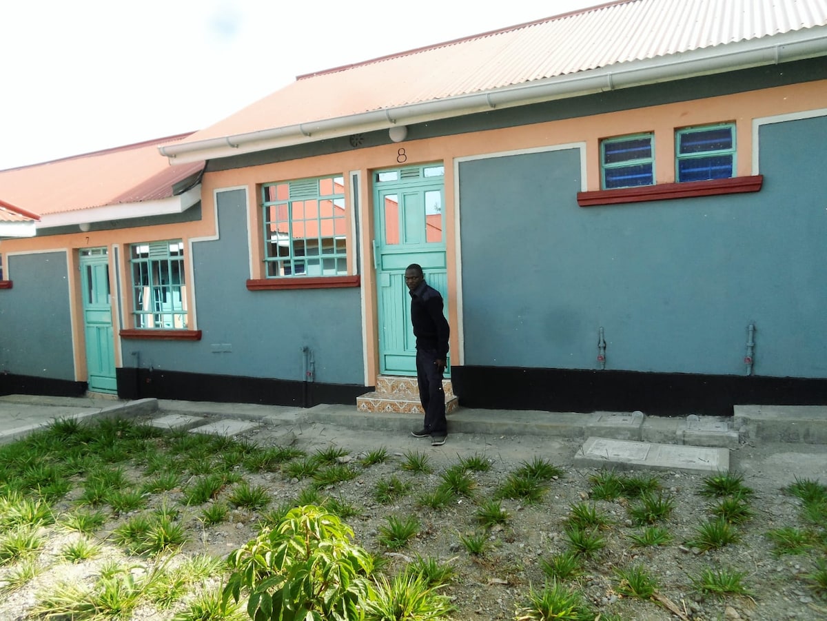 A single-story building with a vibrant exterior is shown, featuring a blue and orange color scheme. The entrance door is painted in a bright teal, framed by multiple windows. A well-maintained area of green plants leads to the structure, enhancing the approachable facade.