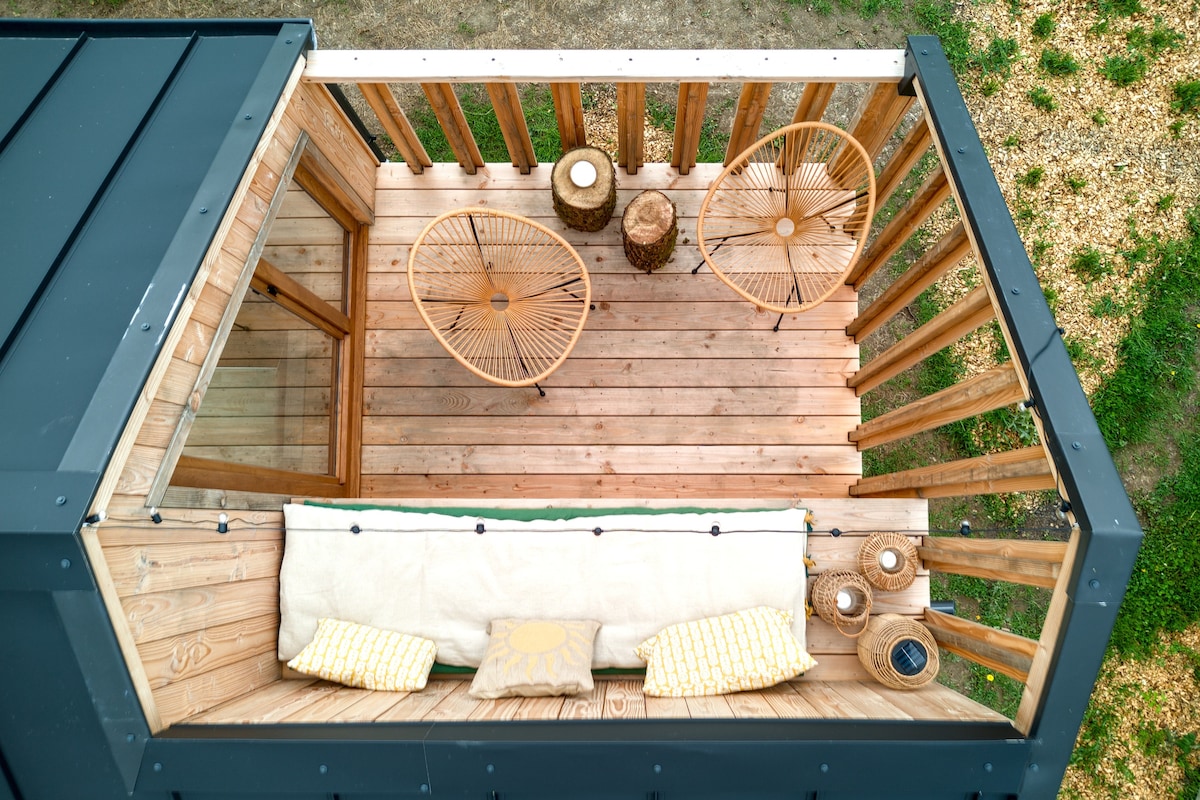 The rooftop terrace features wooden flooring with a cozy seating area. Two chairs made of natural materials and side tables are visible, surrounded by a railing that allows for open views of the surrounding nature.