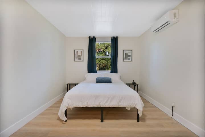 This airy white bedroom features a plush queen bed set beneath a window that fills the space with gentle natural light, great for quiet mornings and restful nights in a calm, minimalist retreat.
