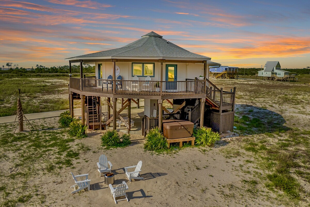 The exterior of the round beach house is highlighted against a vibrant sunset. It features a spacious deck with seating and a hot tub below, surrounded by sand and greenery. A pathway leads from the house towards the beach area, emphasizing its coastal setting.