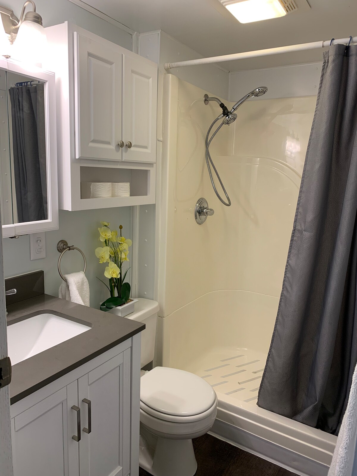 A bright bathroom is equipped with modern fixtures and a spacious shower. The vanity features a sleek countertop and ample storage. A small vase with yellow flowers adds a touch of color, while a gray shower curtain provides privacy.