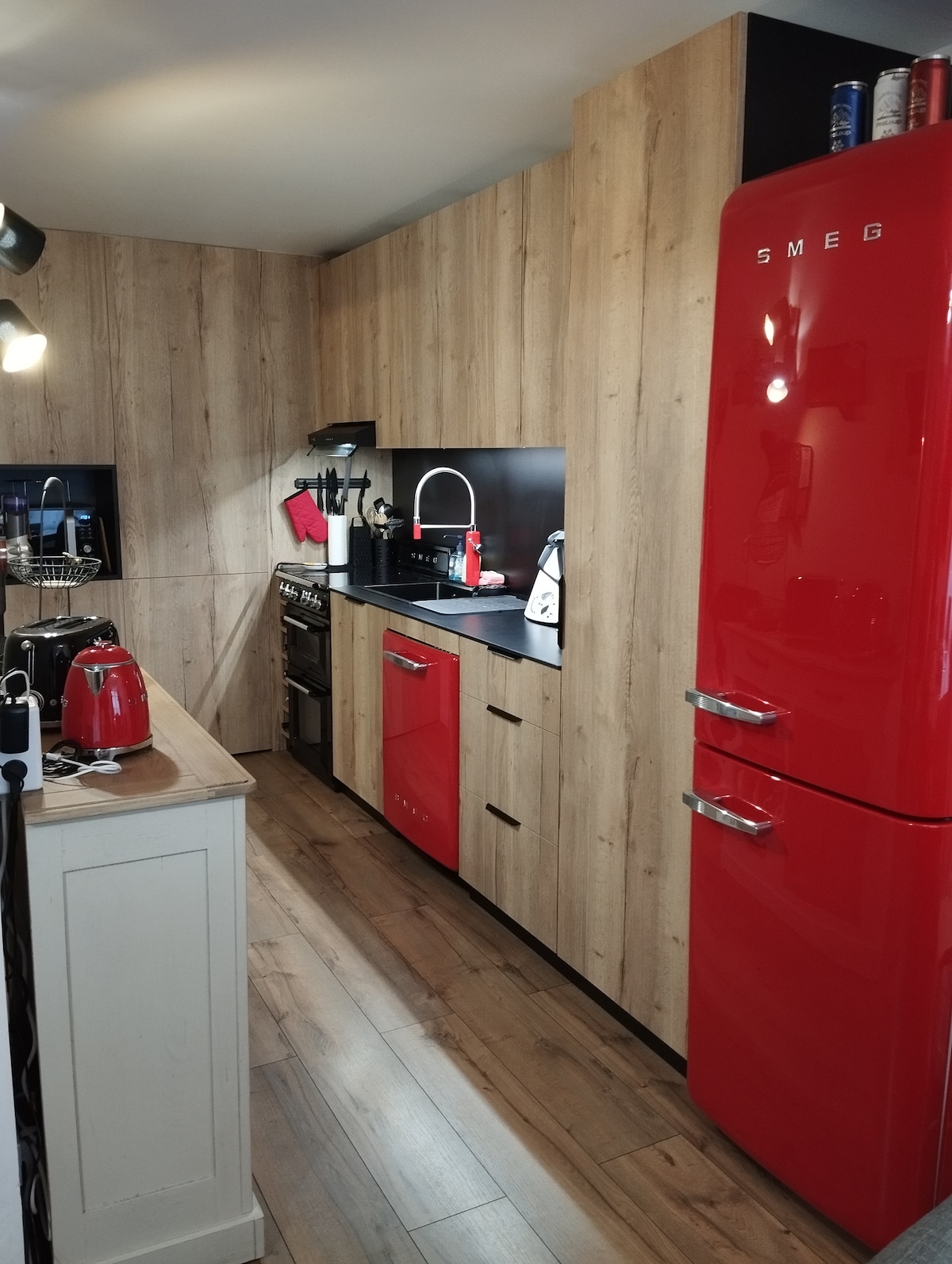 A modern kitchen displays sleek red appliances, including a vintage-style refrigerator, within a wood-paneled setting. Surfaces are dark and glossy, complementing the vibrant cabinetry. Equipped with a stove and various kitchen utensils, the space offers both functionality and style for meal preparation.