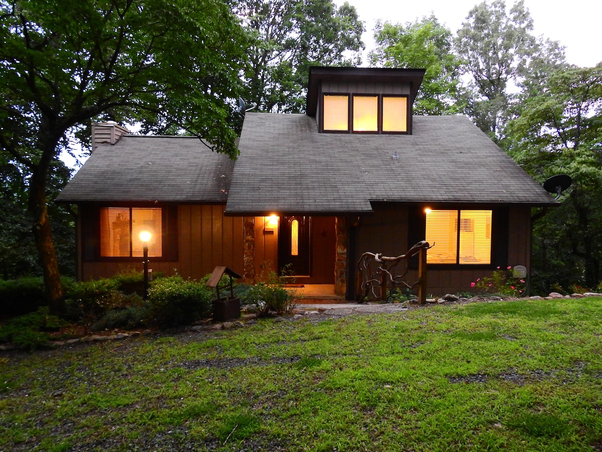 The exterior of Cozy Bear Lodge presents a spacious mountain house, featuring a gabled roof and warm glowing lights from the windows. Surrounded by trees, the path leading to the entrance is framed by greenery, contributing to a serene and inviting atmosphere.