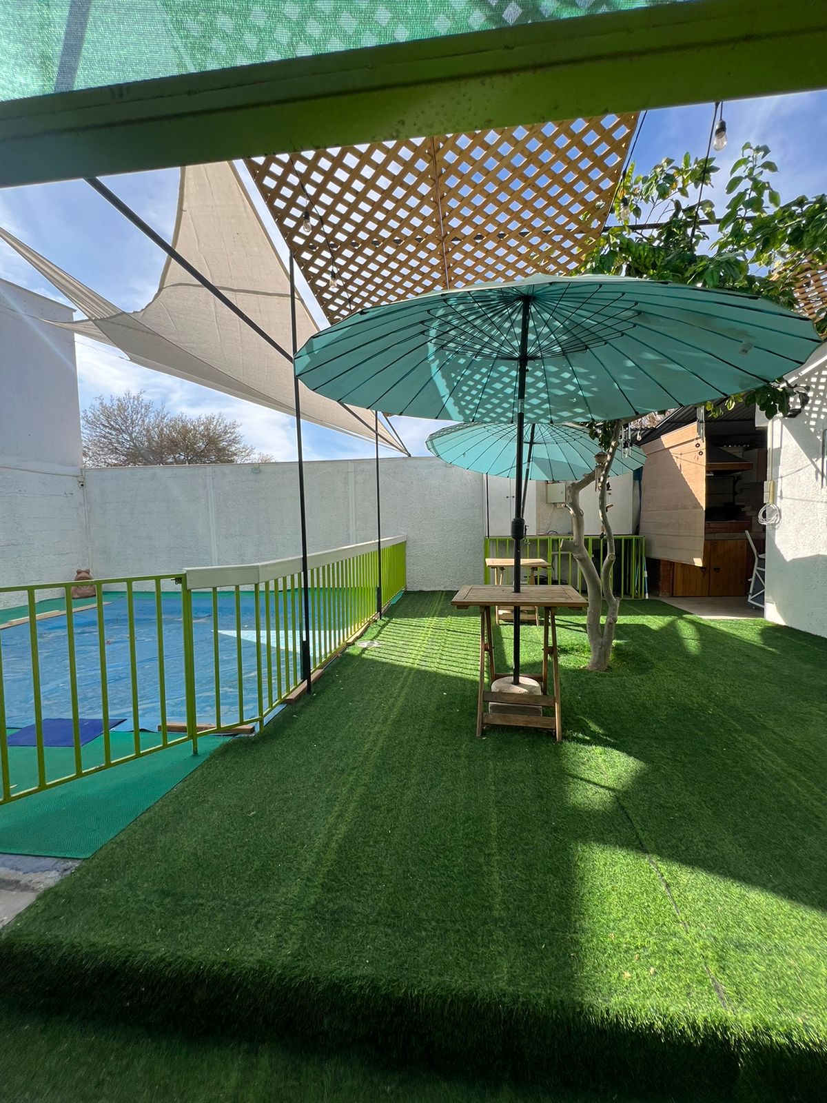 An outdoor area features a swimming pool surrounded by green grass-like turf. Two blue umbrellas provide shade over wooden tables, while a fabric canopy offers additional protection from the sun. The enclosed space is brightened by natural light, creating a relaxed atmosphere.