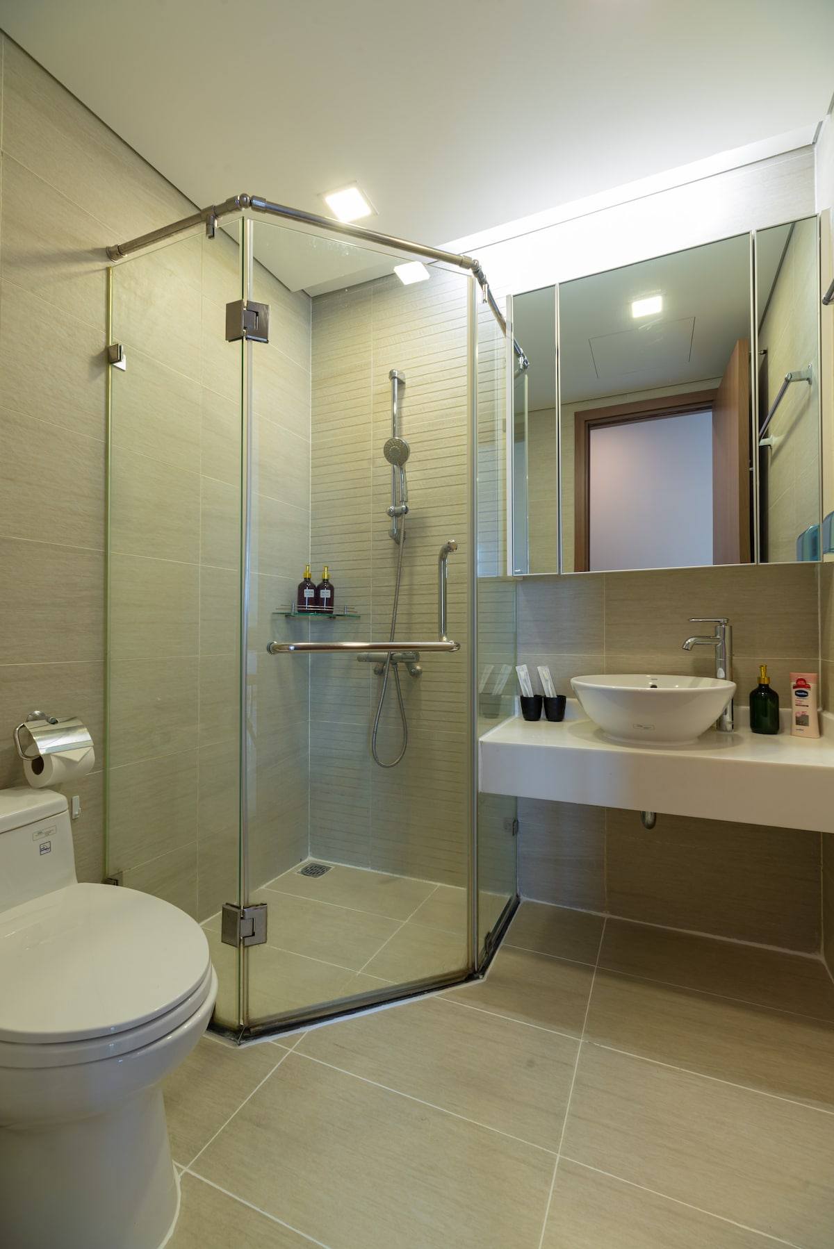 A modern bathroom is equipped with a glass-enclosed shower featuring a rain showerhead and adjustable handheld fixture. A sleek countertop holds a round vessel sink, with toiletries neatly arranged on display. A toilet is positioned adjacent to the shower, with neutral tiles complementing the overall design.