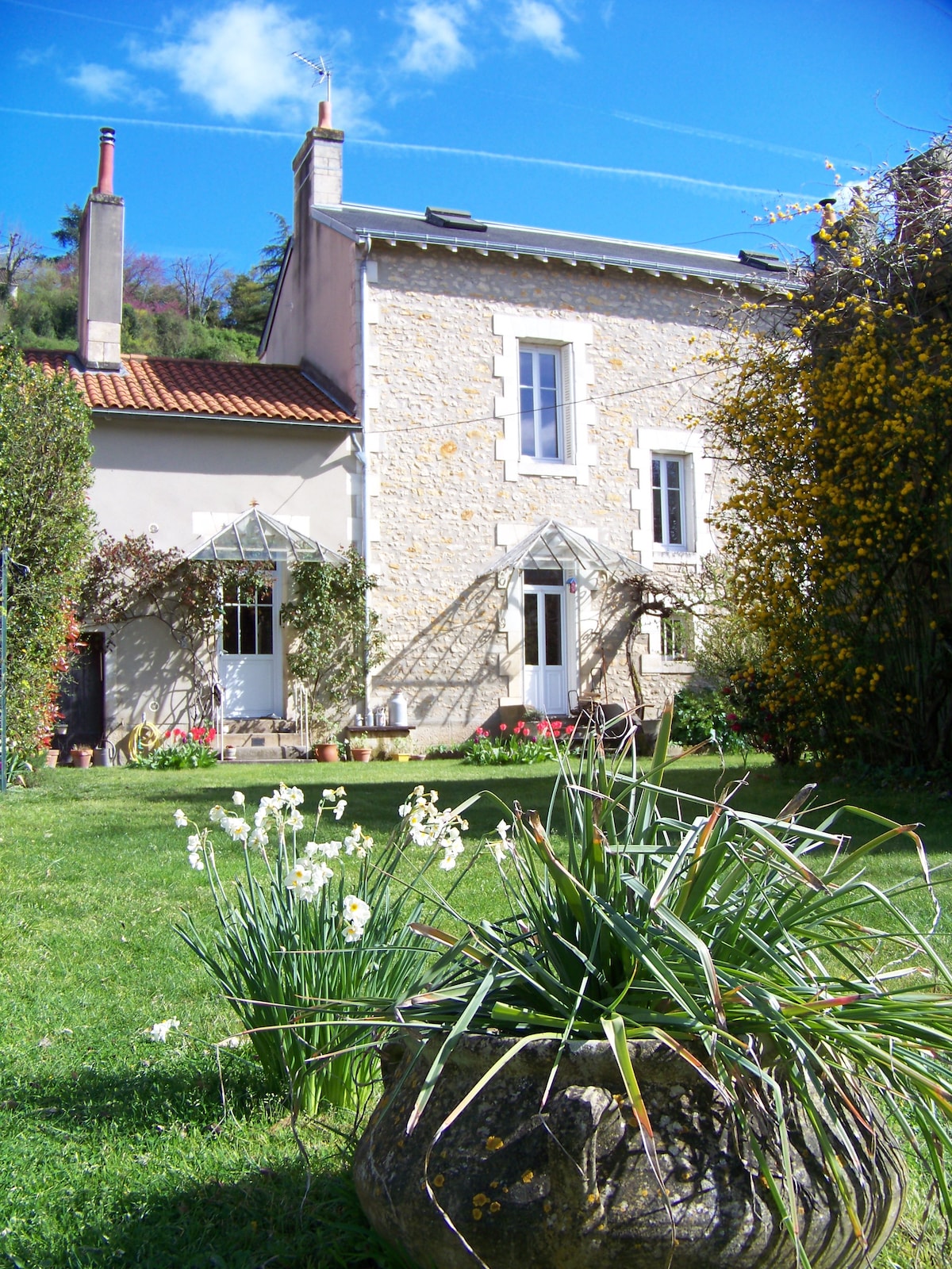 A quaint stone house is surrounded by a lush green garden, highlighting vibrant flora. The structure features white window frames and elegant glass panels. Sunshine filters through, creating a serene setting with clear blue skies and a few clouds above.
