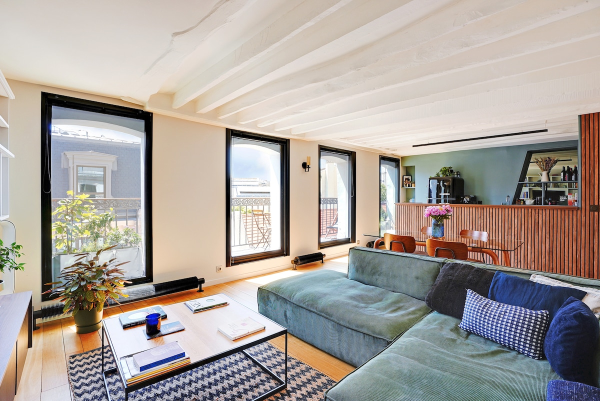 A spacious living area is highlighted by large windows allowing natural light to fill the space. A comfortable sectional sofa faces a stylish wooden bar counter. A small table is positioned in front of the sofa, complemented by plants and decorative items enhancing the inviting environment.
