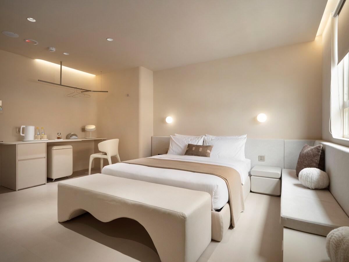 A minimalistic guest room features a spacious bed dressed in neutral tones, complemented by a cozy bench at its foot. Soft lighting illuminates the room, while a desk and a chair provide a work area. A window offers natural light, enhancing the serene environment.