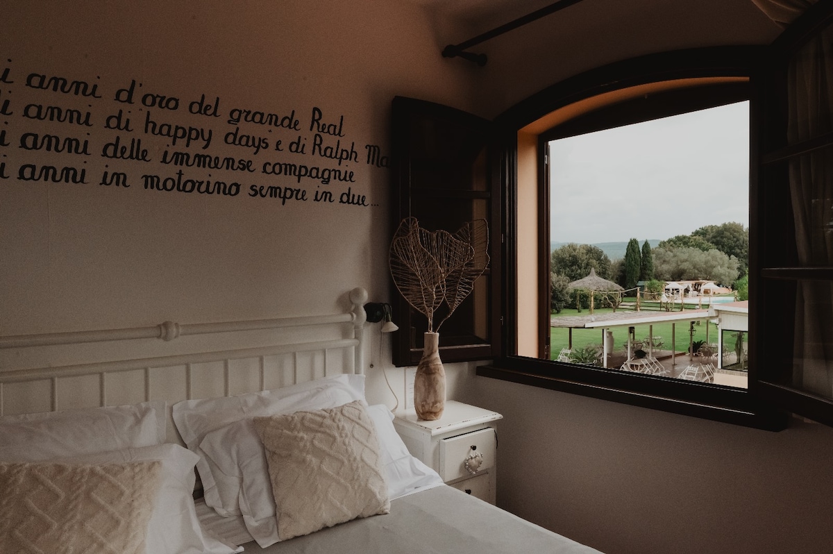 A charming bedroom features a large window overlooking a scenic outdoor area. Light-colored bedding is neatly arranged on the bed, while a decorative vase sits on the nightstand beside it. A calming wall inscription adds a personal touch to the space.