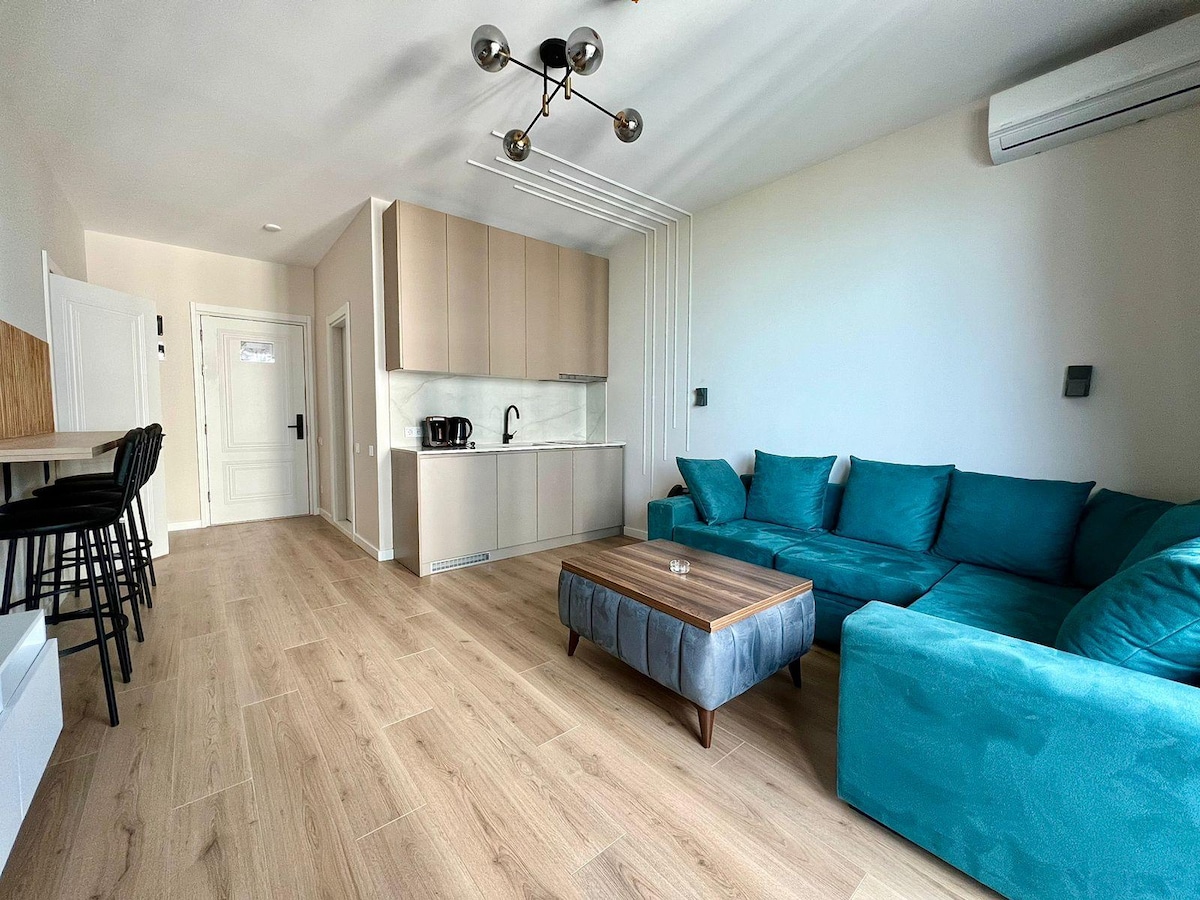 A modern living space features a spacious turquoise sofa paired with a wooden coffee table, set on light-colored flooring. The kitchenette showcases sleek cabinets, a sink, and essential appliances. Bar stools are positioned at a breakfast counter, and natural light fills the room.