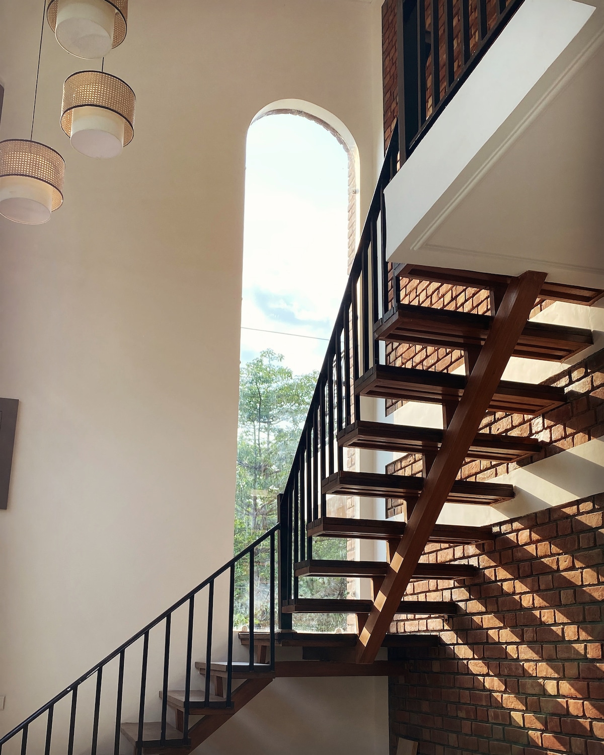 A modern staircase made of polished wood and metal railings ascends alongside a large arched window. Natural light is allowed to filter in, highlighting the textured brick wall and creating an airy ambiance in the space.