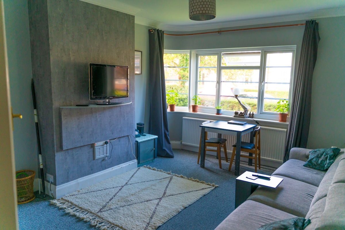 A light-filled living space features a wall-mounted television and a small dining table with two chairs. A comfortable sofa is positioned nearby, and large windows invite natural light, showcasing views of greenery outside. Soft gray curtains frame the windows.