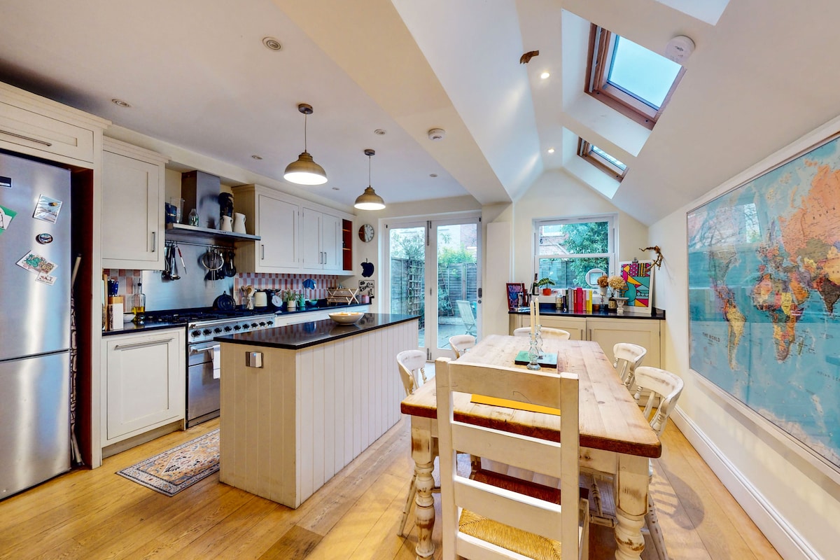 The kitchen features an open layout with modern appliances and a spacious island counter. A dining table with white chairs is positioned centrally, while skylights and large windows enhance the bright ambiance. Colorful wall art adds a touch of character to the space.