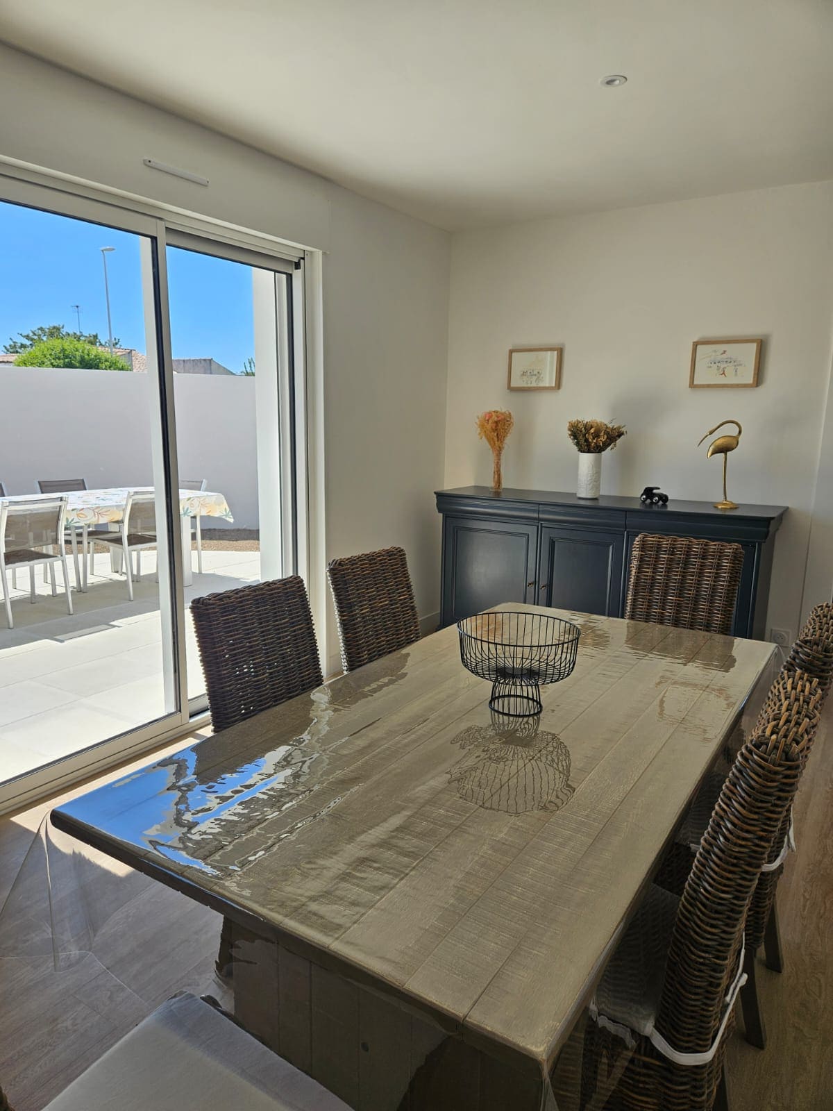 A dining area is presented with a simple wooden table surrounded by six woven chairs. A vase with decorative elements sits in the center. Natural light flows through a sliding glass door, which leads to an outdoor area visible beyond.