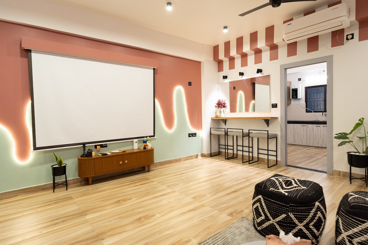 A spacious living area features a large projection screen mounted on a wall with a colorful design. Two bar-style chairs are positioned near a mirrored area. Soft lighting enhances the wood flooring, while plants and decorative elements add a touch of vibrancy.