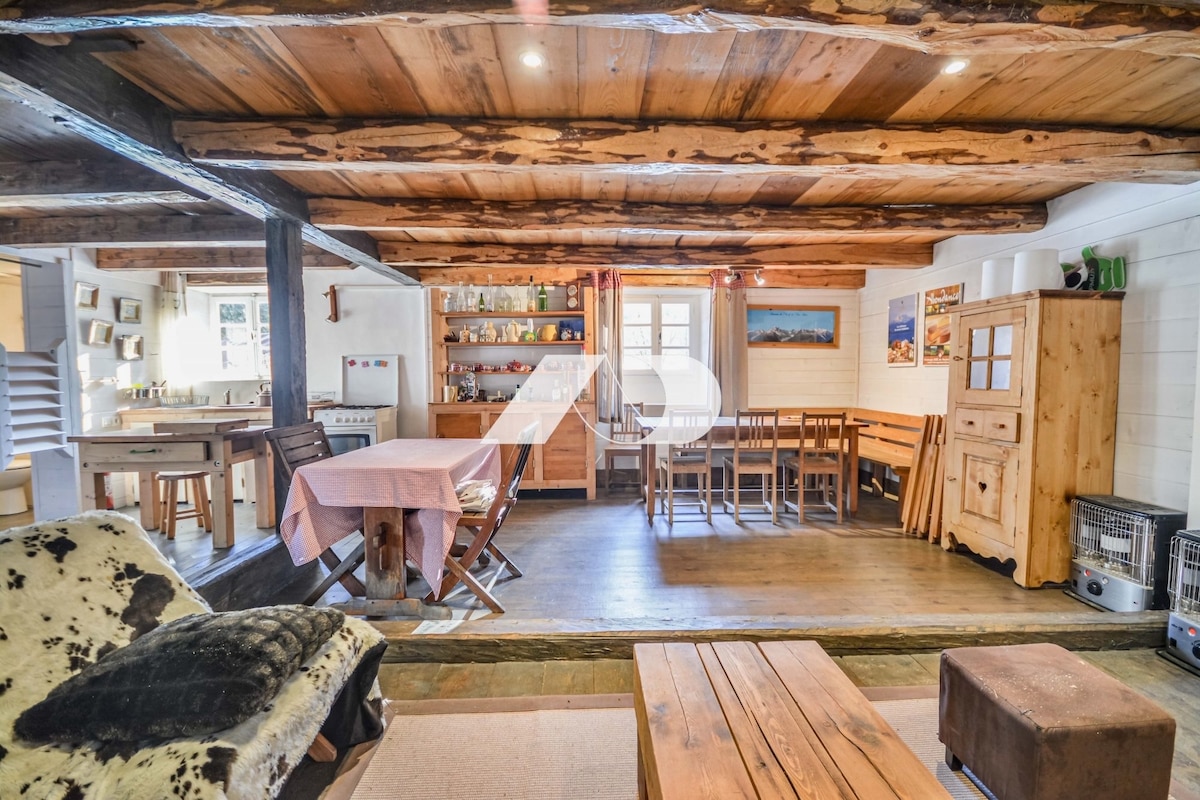An open-plan living space features wooden beams and rustic charm. A large dining table is positioned near the kitchen, while a cozy seating area is visible, complete with a coffee table and a wood-burning stove. Natural light fills the room from multiple windows.