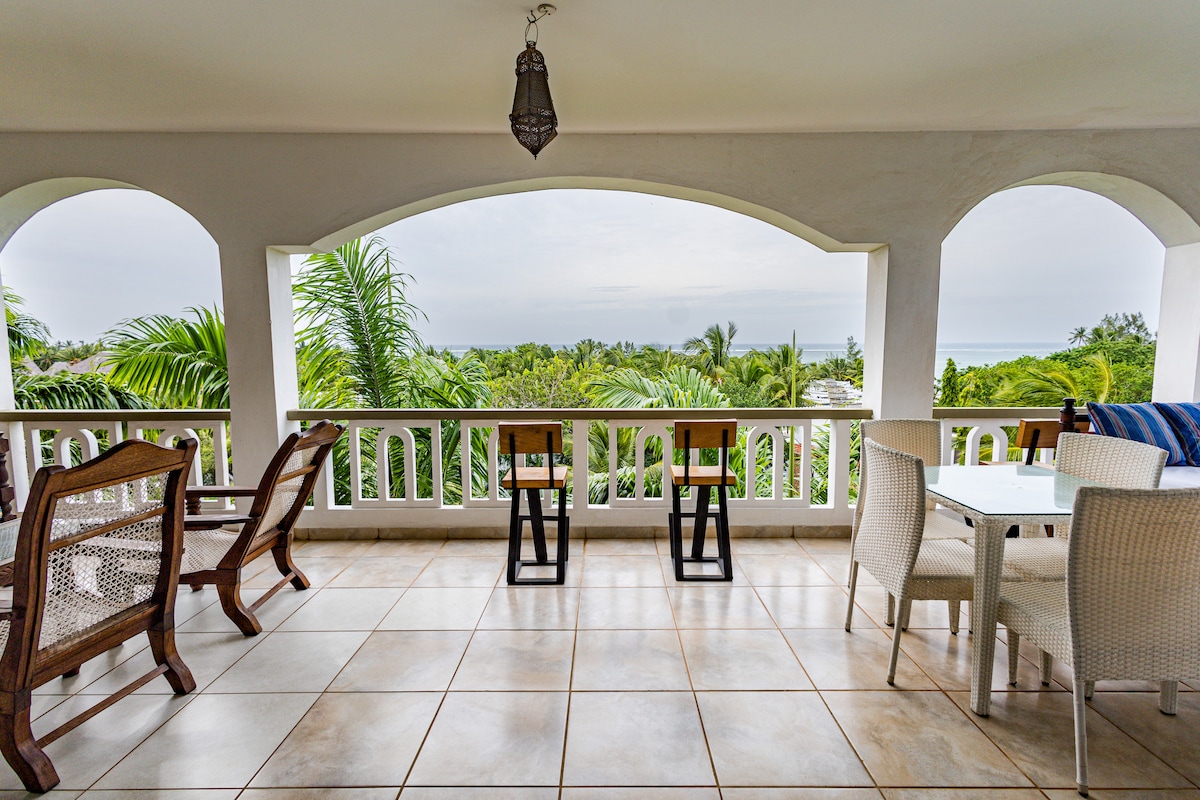 An airy balcony offers sweeping views of lush greenery and the ocean beyond. The space features a combination of wooden and wicker furniture, including two high chairs and a dining table surrounded by comfortable seating. Sunlight gently illuminates the tiled floor.