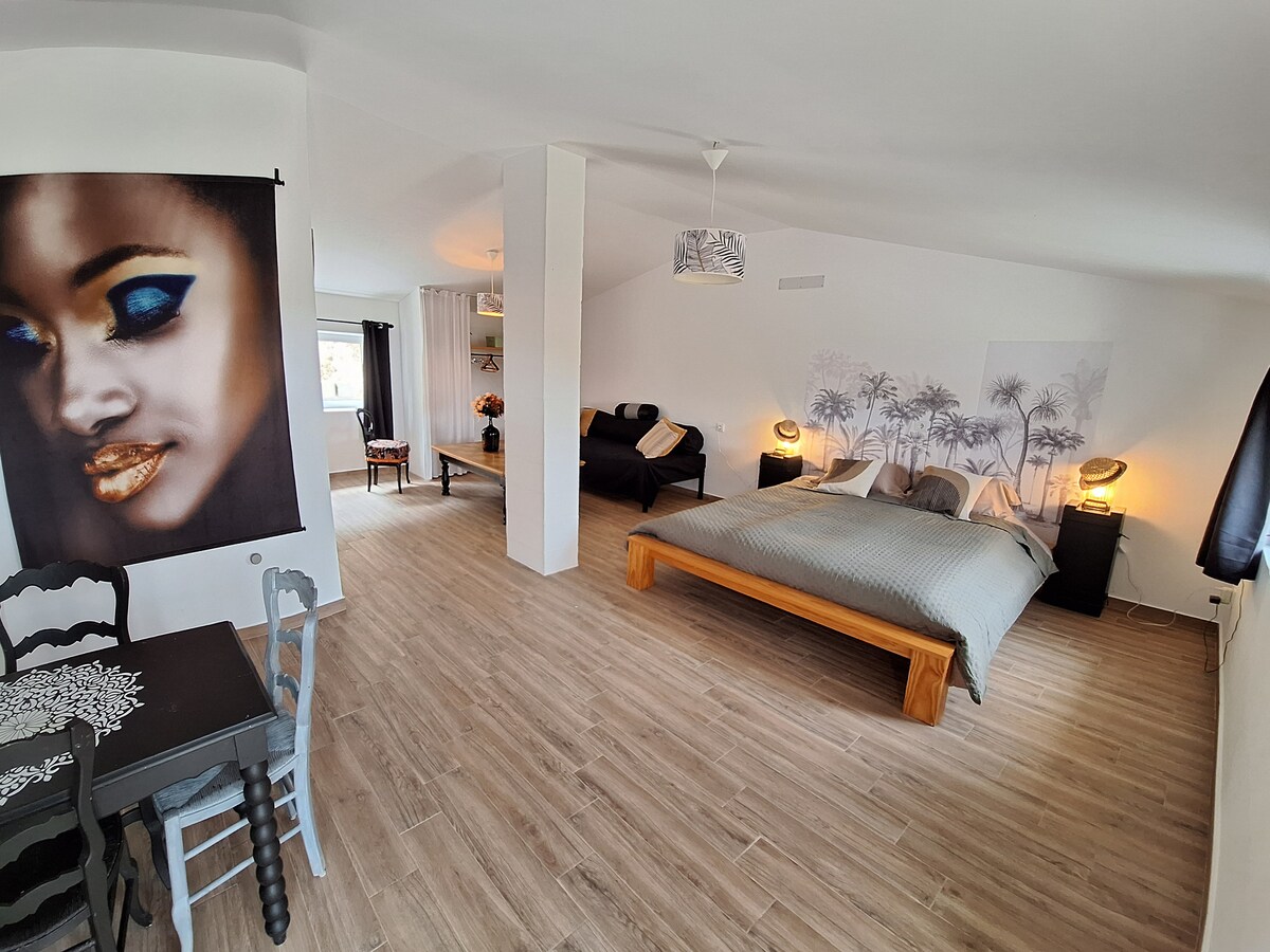 A spacious studio interior features a wooden bed with a neutral-colored bedspread, surrounded by natural light from nearby windows. A cozy seating area is present with dark cushions, and a small dining table with chairs adds functionality to the space. Light-colored wood flooring completes the aesthetic.