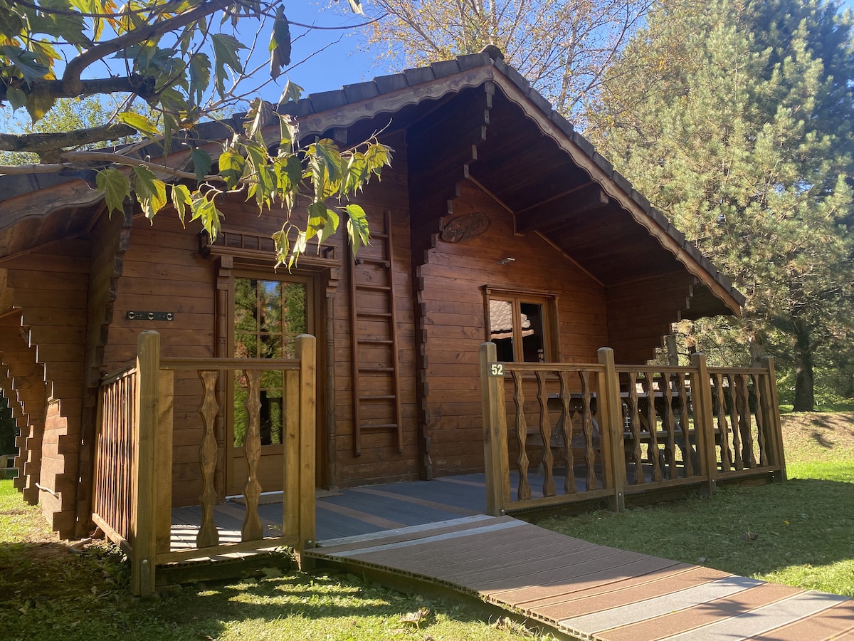 A wooden cabin with a sloped roof and large front porch is surrounded by nature. The entrance is marked by a welcoming pathway and features wooden railing. Sunlight filters through the trees, highlighting the natural beauty of the surroundings.