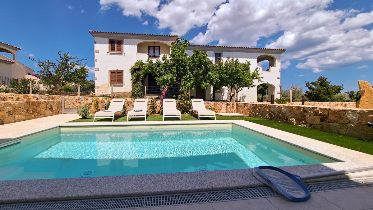 A serene outdoor area showcases a private swimming pool surrounded by five white loungers. Lush greenery and stone walls provide a calming backdrop, with a multi-story residential building visible in the background against a bright blue sky dotted with clouds.