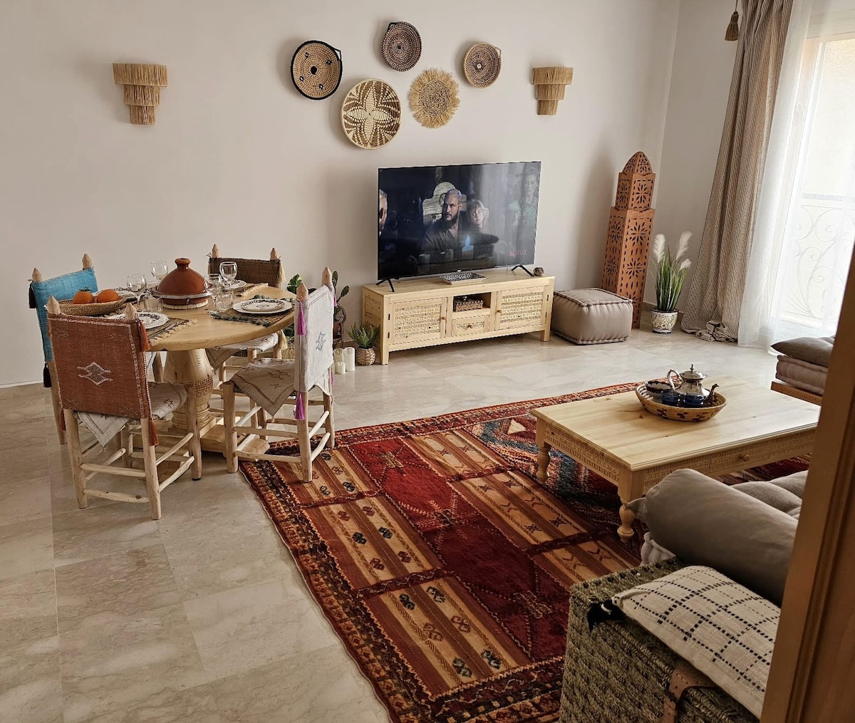 A cozy living area features a light-colored marble floor and a patterned rug. A wooden coffee table and a comfortable seating arrangement are complemented by decorative elements. A Smart TV is positioned against a wall adorned with artisanal decor, and a dining table awaits guests with dinnerware set.