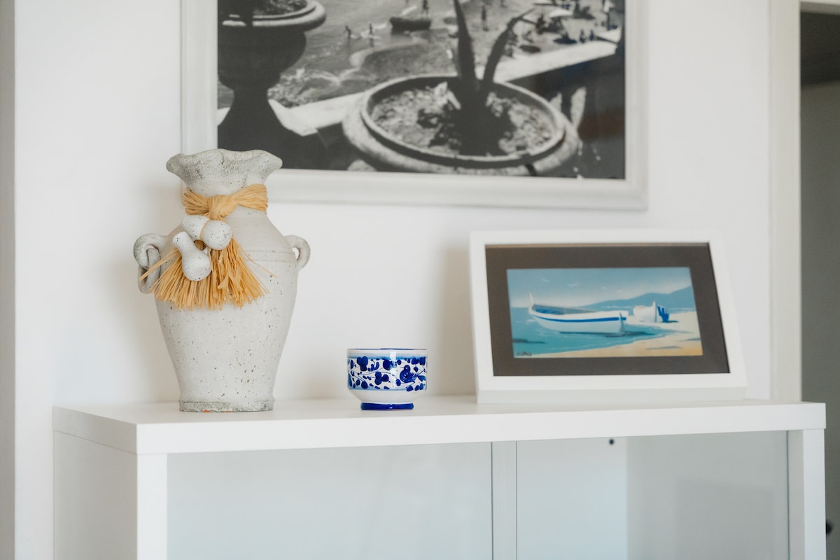A decorative display features a grey ceramic vase with a tassel and small embellishments, positioned beside a blue and white patterned bowl. An art print depicting a coastal scene rests in a frame on the table, while a black-and-white photograph is displayed in the background.