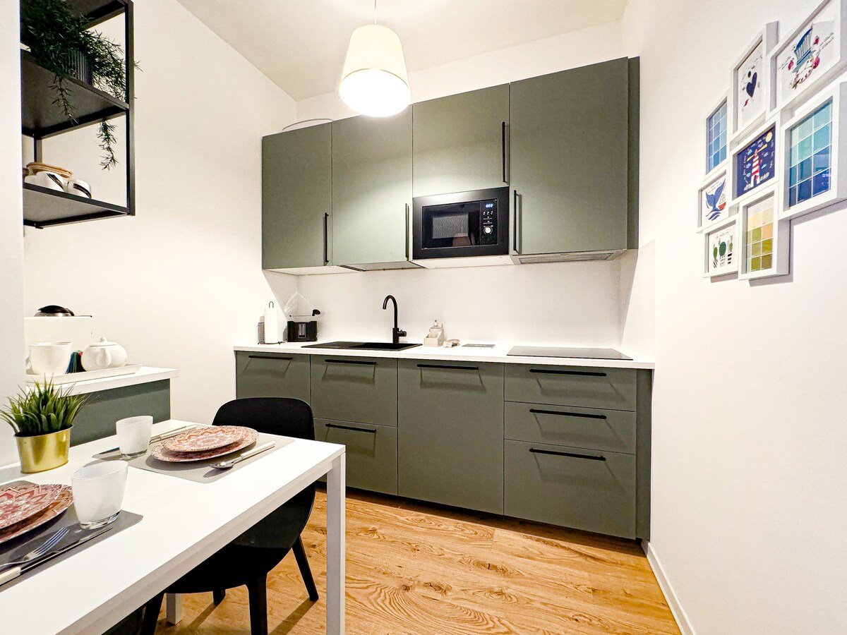 A modern kitchen features sleek green cabinetry and a white countertop. Amenities include a microwave and a kettle. A dining table with chairs is set for meals, with plates and utensils arranged neatly. Wall art in various colors adds a decorative touch to the space.