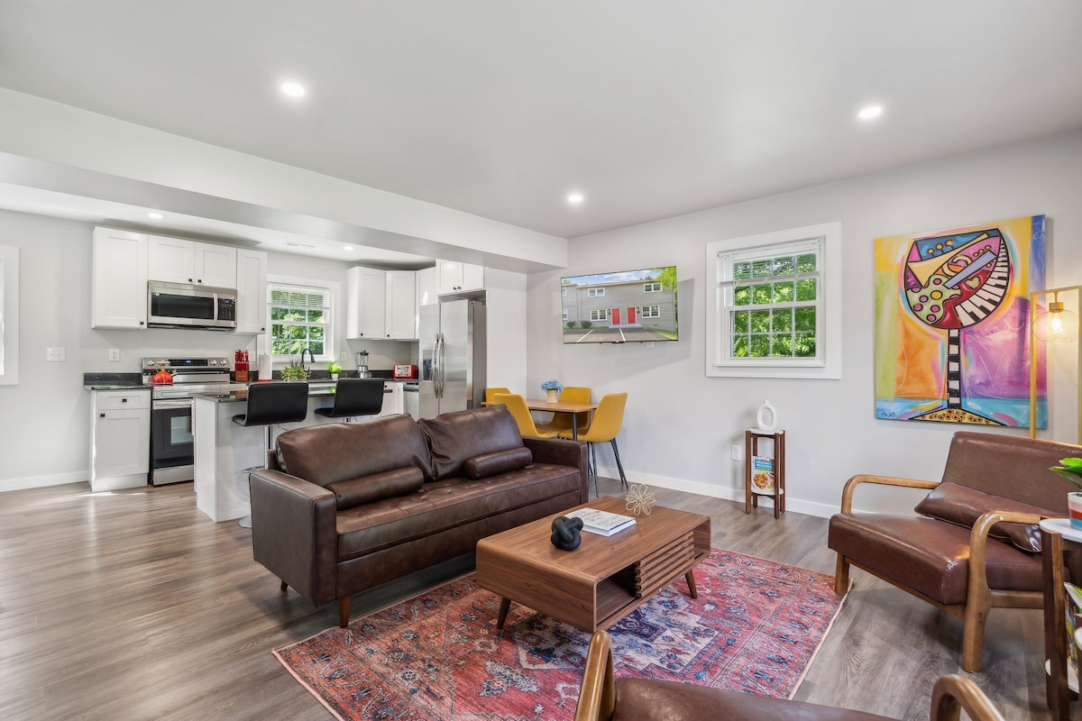 A modern living area features a brown leather sofa and two armchairs, paired with a wooden coffee table on a patterned rug. Open kitchen and dining space are visible, showcasing stainless steel appliances and a colorful art piece on the wall.
