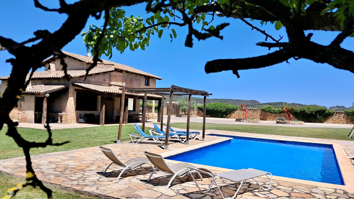 A private garden setting features a rectangular swimming pool surrounded by a stone patio. Sun loungers are arranged nearby under a shaded structure, and spacious seating areas are visible in the background. Lush greenery complements the tranquil outdoor environment.